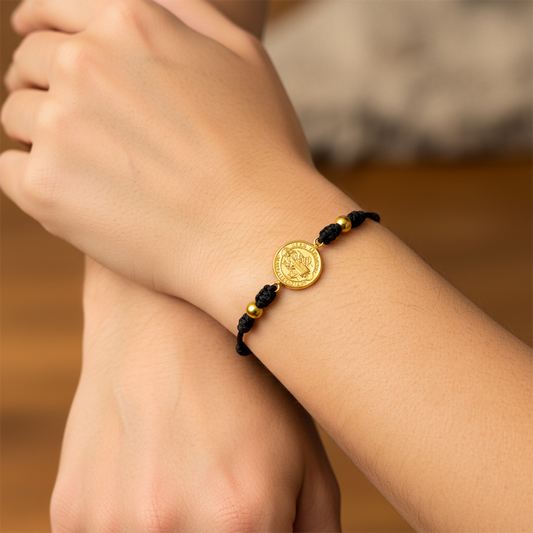 Mano de modelo usando una pulsera tejida en hilo color negro con dije San Benito y esferas lisas en oro amarillo de 18 quilates de Joyería Caracas. 