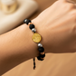 Mano de modelo usando una pulsera tejida en hilo color negro, con esferas lisas y facetadas en 3 oros y dije de San Benito de 18 quilates de Joyería Caracas. 