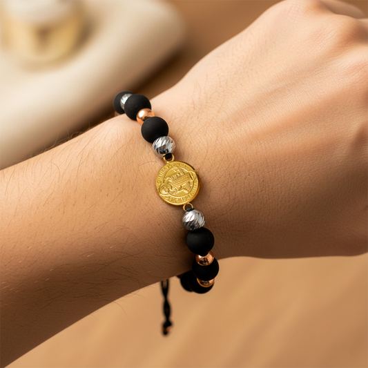 Mano de modelo usando una pulsera tejida en hilo color negro, con esferas lisas y facetadas en 3 oros y dije de San Benito de 18 quilates de Joyería Caracas. 
