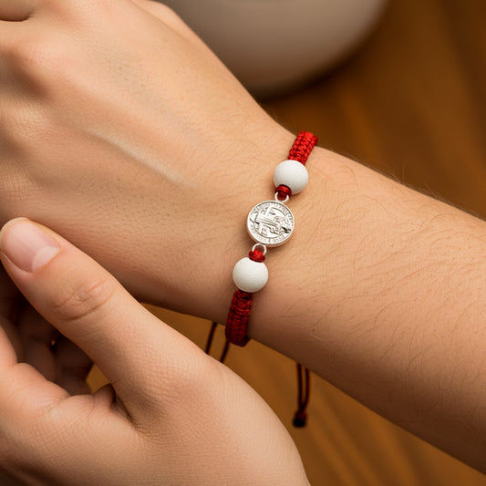 Mano de modelo usando una pulsera tejida en hilo de color rojo con dije de San Benito en oro blanco de 18 quilates con bisutería de calidad de Joyería Caracas. 