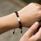 Mano de modelo usando una pulsera tejida en hilo de color vinotinto con dije de San Benito en oro blanco de 18 quilates con bisutería de calidad de Joyería Caracas. 