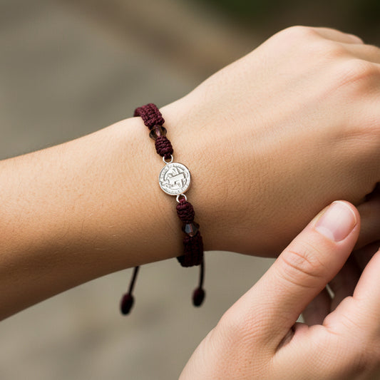 Mano de modelo usando una pulsera tejida en hilo de color vinotinto con dije de San Benito en oro blanco de 18 quilates con bisutería de calidad de Joyería Caracas. 