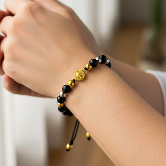 Mano de modelo usando una pulsera tejida en hilo color negro con dije San Benito y esferas lisas en oro amarillo y blanco de 18 quilates con bisutería de calidad de Joyería Caracas. 