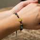 Mano de modelo usando una pulsera tejida en hilo color negro con dije de San Benito y 2 esferas lisas en oro amarillo de 18 quilates y bisutería de calidad de Joyería Caracas. 