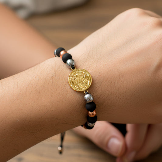 Mano de modelo usando una pulsera tejida en hilo color negro con dije de San Benito en oro amarillo y esferas lisas en oro blanco y rosa de 18 quilates de Joyería Caracas. 