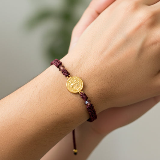 Mano de modelo usando una pulsera tejida en hilo color vinotinto con dije de San Benito en oro amarillo de 18 quilates de Joyería Caracas. 
