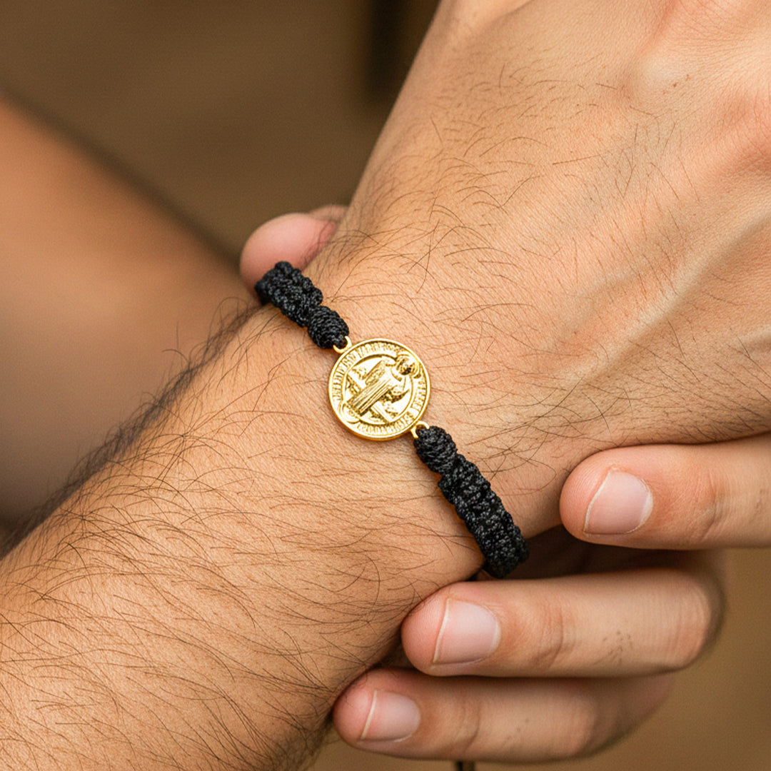 Mano de hombre usando una pulsera tejida con herraje de San Benito en oro amarillo de 18 quilates de Joyería Caracas. 