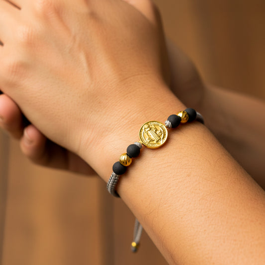 Mano de modelo usando una pulsera tejida con hilo de color gris con dijes de San Benito XL y esferas facetadas en oro amarillo de 18 quilates con bisutería de Joyería Caracas. 