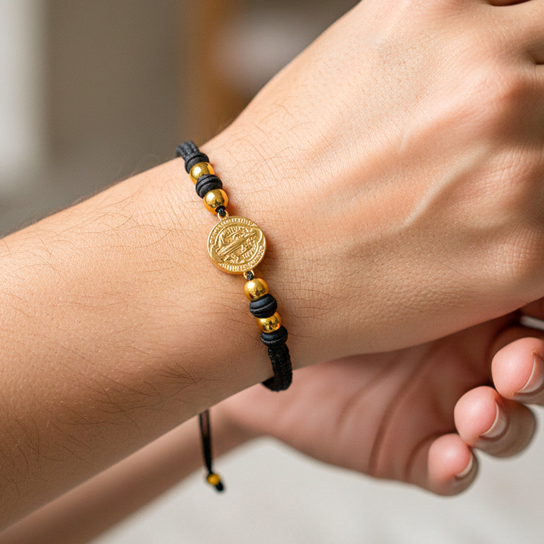 Mano de modelo usando una pulsera tejida en hilo color negro con accesorios, herraje de San Benito con esferas lisas en oro amarillo de 18 quilates de Joyería Caracas. 