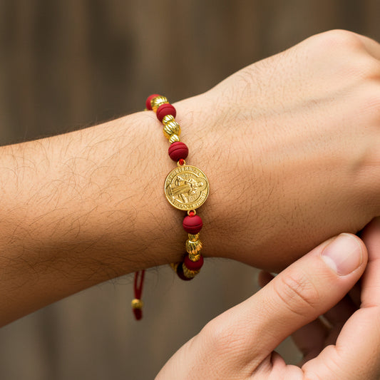 Mano de modelo usando una pulsera tejida en hilo de color rojo, con esferas facetadas y herraje con forma de San Benito en oro amarillo de 18 quilates con bisutería de calidad de Joyería Caracas. 