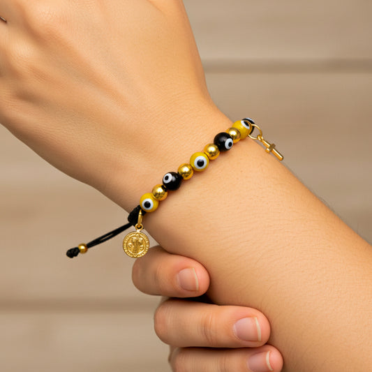 Mano de modelo usando una pulsera tejida en hilo color negro, con dije en forma de San Benito, cruz y esferas lisas en oro amarillo de 18 quilates con bisutería de calidad de Joyería Caracas. 
