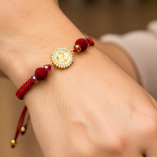 Mano de modelo usando una pulsera tejida en hilo color rojo con dije de San Benito en oro amarillo de 18 quilates con cristales de color blanco de Joyería Caracas. 
