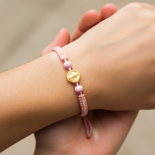 Mano de modelo usando una pulsera tejida en hilo de color rosa con dije de San Benito en oro amarillo de 18 quilates con bisutería de calidad de Joyería Caracas. 