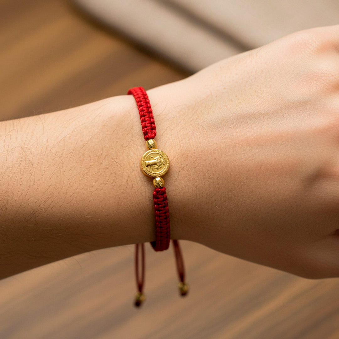 Mano de modelo usando una pulsera tejida en hilo de color rojo con dije de San Benito y 2 esferas facetadas en oro amarillo de 18 quilates de Joyería Caracas. 
