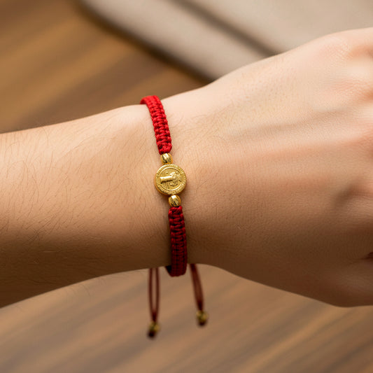 Mano de modelo usando una pulsera tejida en hilo de color rojo con dije de San Benito y 2 esferas facetadas en oro amarillo de 18 quilates de Joyería Caracas. 