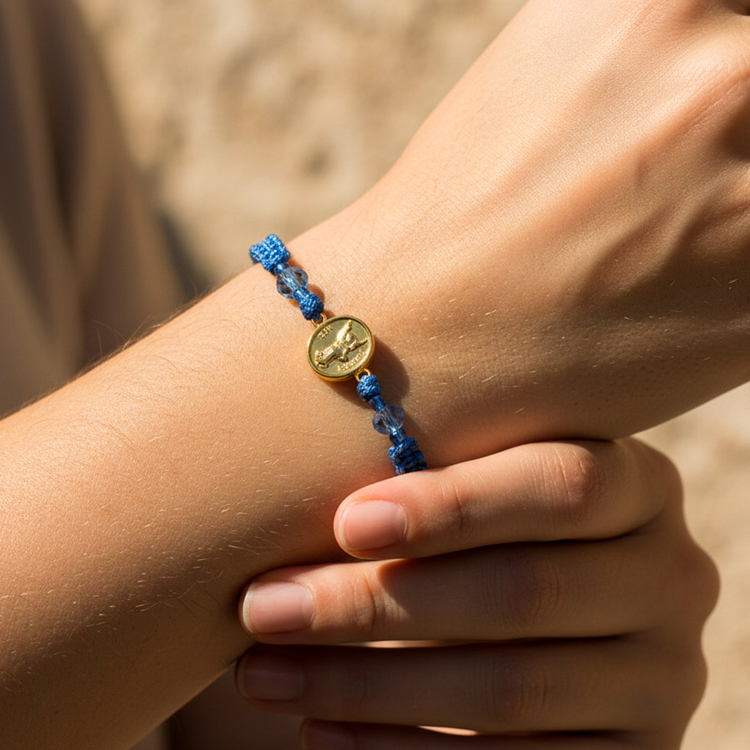 Mano de modelo usando una pulsera tejida en hilo color celeste con dije del Arcángel San Gabriel en oro amarillo de 18 quilates con bisutería de calidad de Joyería Caracas.