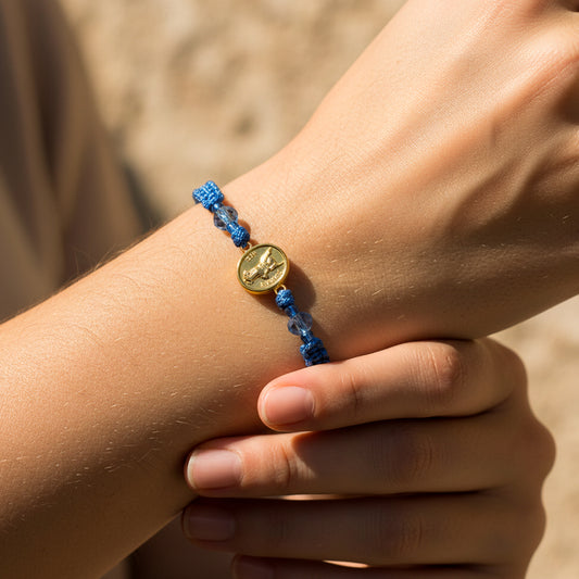 Mano de modelo usando una pulsera tejida en hilo color celeste con dije del Arcángel San Gabriel en oro amarillo de 18 quilates con bisutería de calidad de Joyería Caracas.