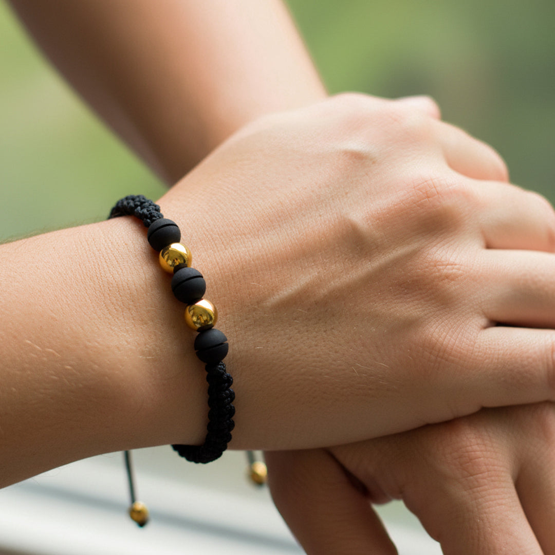 Mano de modelo usando una pulsera tejida en hilo color negro con 2 esferas lisas en oro amarillo de 18 quilates y bisutería de calidad de Joyería Caracas. 