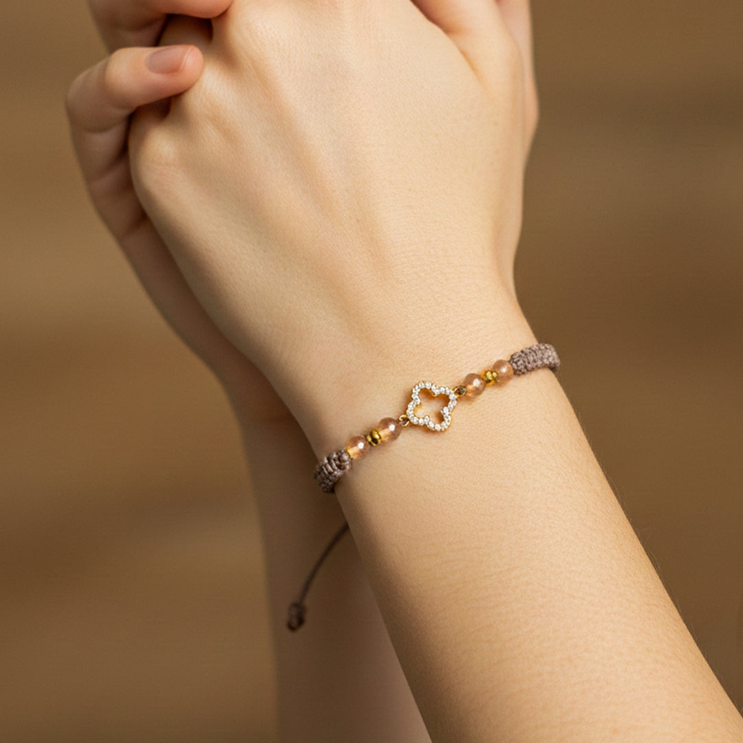 Mano de modelo usando una pulsera tejida en hilo color café con accesorios, herraje en forma de trébol con cristales y esferas de Joyería Caracas. 